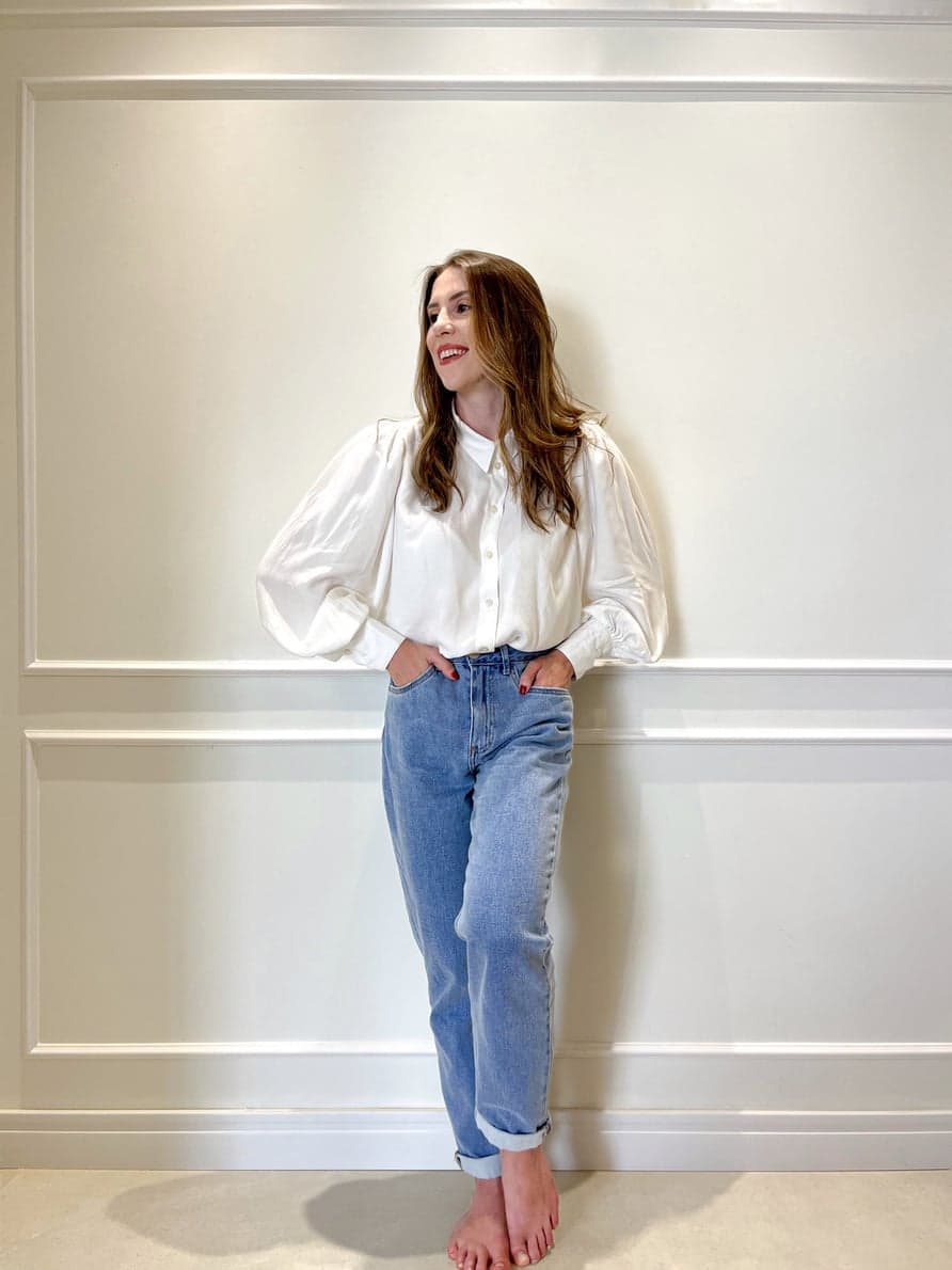 Mariana Coppini de camisa branca e calça jeans, apoiada em uma parede clara